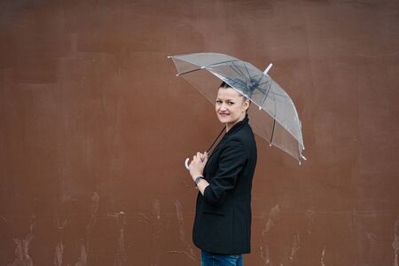 A young attractive woman in a black jacket and blue jeans posing outdoor against the background of the building. Holds a transparent umbrella. Rainy weather.の写真素材