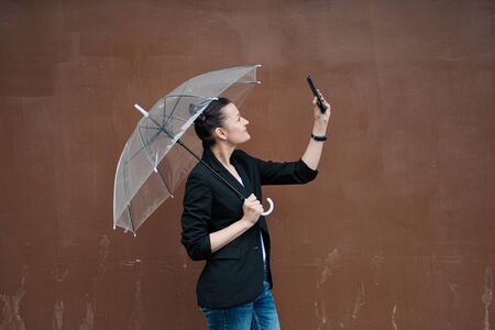 A young attractive woman in a black jacket and blue jeans posing outdoor against the background of the building. Holds a transparent umbrella. Rainy weather.の写真素材