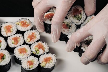 Cooks hands close-up. A male chef makes sushi and rolls from rice, red fish and avocado. White gloves. Dark background.の写真素材