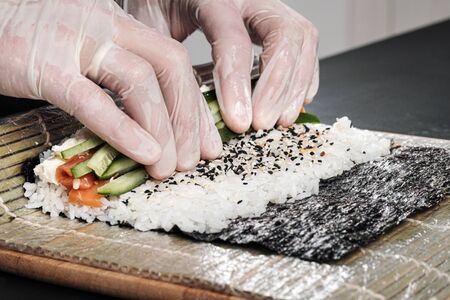 Cooks hands close-up. A male chef makes sushi and rolls from rice, red fish and avocado. White gloves. Dark background.の写真素材