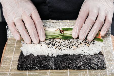 Cooks hands close-up. A male chef makes sushi and rolls from rice, red fish and avocado. White gloves. Dark background.の写真素材