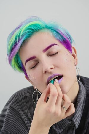A young woman with short pixie bob haircut and dyed hair, holds in the mouth and in the hands gemstones and jewels. White background in a bright room.の写真素材