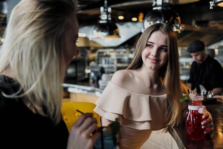 Two young smart beautiful girls in dresses are sitting at the bar. Drink red and blue cocktails from glasses with tubules, communicate and laugh.の写真素材
