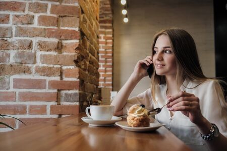 A young beautiful brunette girl in a loft-style cafe sits at a table by the window, drinks coffee and uses a smartphone, calls, takes pictures and types a message.の写真素材