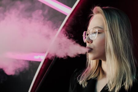 Two young smart beautiful girls in dresses sit at a table in a nightclub or hookah. They smoke hookah and smoke and drink cocktails from glasses with tubules, talk and laugh. Take a selfie on the phone.の写真素材