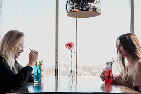 Two young smart beautiful girls in dresses sit at a restaurant table opposite a large window. Drink red and blue cocktails from glasses with tubules, communicate and laugh.の写真素材