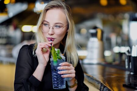 A young beautiful blond woman with glasses sits at the bar in a yellow interior. Drinks alcoholic cocktail with a straw and green mint. Night life. and rest at a party.の写真素材