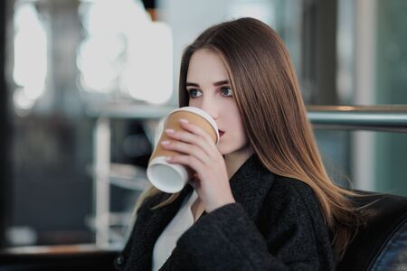 A young beautiful brunette girl in a gray coat is sitting on a bench waiting. Drinking coffee and holding a paper cup with a drink. Gray blurred background.の写真素材