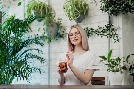 Young beautiful athletic woman blonde in glasses sits at a table in a green cafe and drinks strawberry lemonade from a glass jar. Smiles and laughs.の写真素材