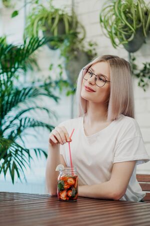 Young beautiful athletic woman blonde in glasses sits at a table in a green cafe and drinks strawberry lemonade from a glass jar. Smiles and laughs.の写真素材
