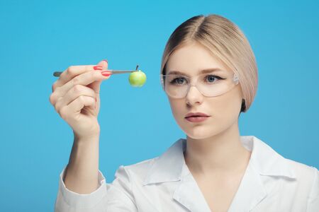 A scientist biologist woman in the laboratory examines a genetically modified green small apple, in a white coat, on a blue background. GMO concept and modification of food.の写真素材