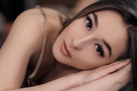 Beautiful brunette girl with brown eyes with bare shoulders lying on the bed. Face close up. Asian and Oriental type of appearance. Skin care.の写真素材