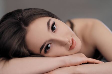 Beautiful brunette girl with brown eyes with bare shoulders lying on the bed. Face close up. Asian...