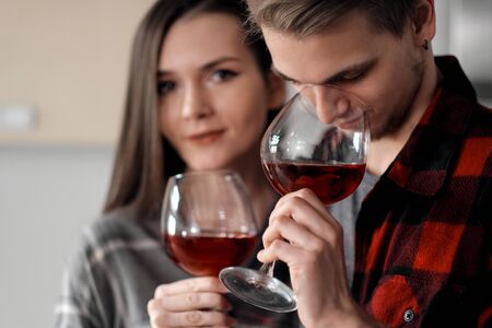 Beautiful young couple in plaid shirts in the kitchen preparing food and drinking red wine from large glasses.の写真素材