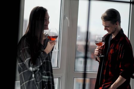 Young beautiful couple in plaid shirts on the balcony of a multistory building. Drink and taste wine, socialize, laugh and embrace.の写真素材