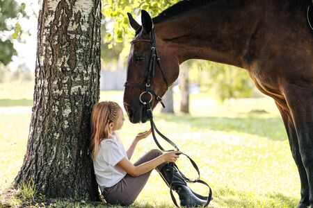 Girl teenager jockey sits in a green clearing under a tree. Feeds a horse an apple and strokes it. Love for horses.の写真素材