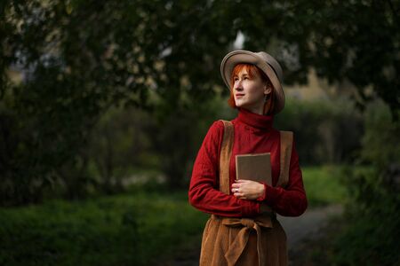 Head portrait of a young attractive girl with red hair in a hat and a red sweater on a background of green trees in the park. She holds a book in hands and reads. Walk outdor.の写真素材