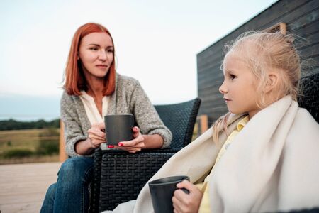 A young mother and little daughter sit in chairs in warm rugs and drink tea from the mugs on the terrace of a wooden house. Autumn evening in the country. The concept of family holidays and relationships.の写真素材