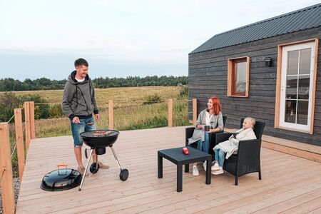 A young Caucasian family couple with a baby daughter sits on the terrace in chairs and drinks hot drinks. Against the background of a small modern rustic wooden house. Father is cooking barbecue. Summer evening in the village.の写真素材
