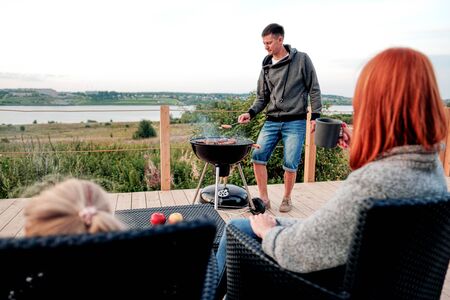 A young Caucasian family couple with a baby daughter sits on the terrace in chairs and drinks hot drinks. Against the background of a small modern rustic wooden house. Father is cooking barbecue. Summer evening in the village.の写真素材