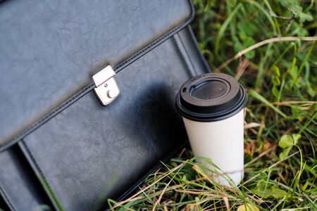 Black business briefcase on the green grass of a park and a paper cup of takeaway coffee. Outdoor...