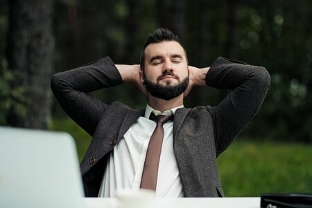 Young attractive male businessman in suit sitting at desk in forest of nature park. Stretches and rests from work. Laptop and cup of coffee on table.の写真素材
