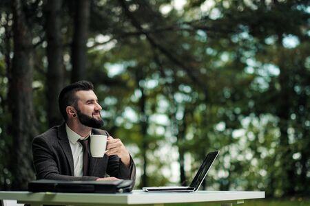 Young attractive business man in suit and tie sits at desk and works on computer outdoors. Drink coffee from white cup. Green trees, nature and park on background. Remote work outside office concept.の写真素材
