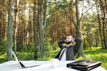 Young attractive male businessman in suit sitting at desk in forest of nature park. Stretches and rests from work. Laptop and cup of coffee on table.の写真素材