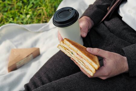 Hands of a man in a suit with a glass of takeaway coffee and a triangular sandwich with tuna. Outdoor lunch and junk food concept.の写真素材