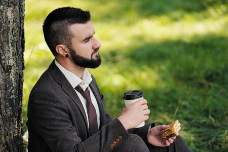 Young attractive bearded business man sitting on green grass under tree and resting in park. Read...