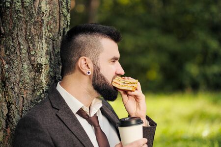 Young attractive business man with beard sitting on green grass under tree and resting in park....