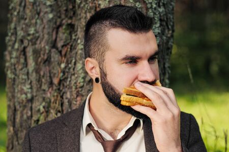 Young attractive business man with beard sitting on green grass under tree and resting in park. Drinks coffee and eats a sandwich. Relaxation, tired of work, lunch break in nature outdoor.の写真素材