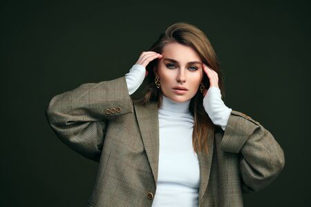 A young beautiful blonde woman in a white turtleneck and a mans jacket poses on a green olive background in the studio. Beauty and elegance.の写真素材