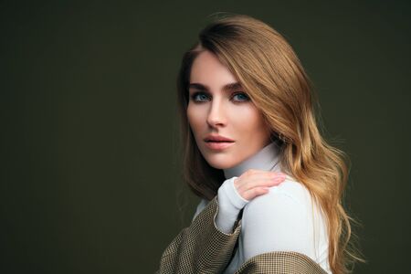 A young beautiful blonde woman in a white turtleneck and a mans jacket poses on a green olive background in the studio. Beauty and elegance.の写真素材