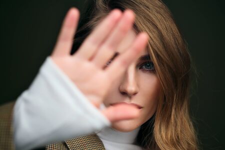 A young beautiful blonde woman in a white turtleneck and a mans jacket poses on a green olive background in the studio and covers face with hand. Beauty and elegance.の写真素材