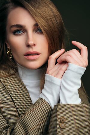 A young beautiful blonde woman in a white turtleneck and a mans jacket poses on a green olive background in the studio. Touches lips with fingers. Beauty and elegance.の写真素材
