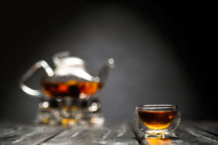 Horizontal photo of the tea set on a black background. Glass transparent teapot and cup. Black leaf tea is brewed.の写真素材