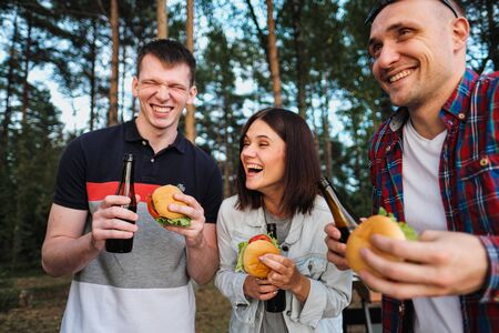 A group of friends rests and relaxes in nature. People eat hamburgers, drink beer, communicate and...