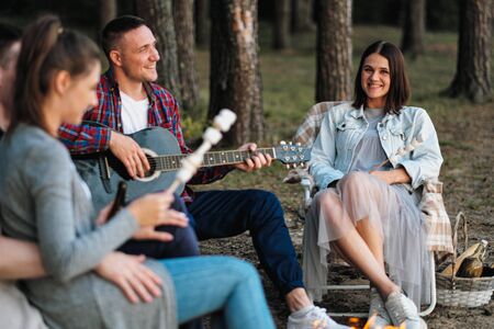 A group of friends relax in a forest camp. Men and women prepare a marshmallow on a bonfire. They talk, smile and laugh. A party in nature.の写真素材