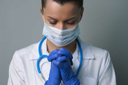 A female doctor therapist in a white robe, mask and gloves. Face close-up. The doctor cries and prays. Tears in eyes. Regret, anxiety and despair. Pandemic and virus epidemic. Coronavirus covid-19.の写真素材