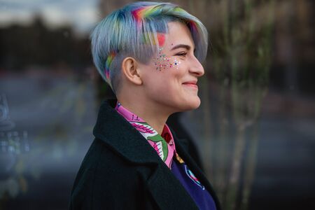 Portrait of a young beautiful woman in green coat with a short haircut and dyed hair. Grey main color and yellow, green, blue and red hair color. Posing in the street.の写真素材
