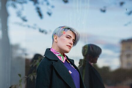 Portrait of a young beautiful woman in green coat with a short haircut and dyed hair. Grey main color and yellow, green, blue and red hair color. Posing in the street.の写真素材
