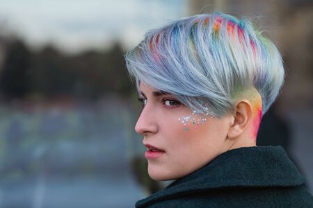 Portrait of a young beautiful woman in green coat with a short haircut and dyed hair. Grey main color and yellow, green, blue and red hair color. Posing in the street.の写真素材