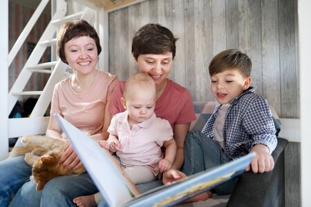 A young lesbian same-sex family with two children, a son and a daughter spend time at home. They sit on the couch and read a book. The pet cat is lying next to it. Homosexual couple marriage.の写真素材