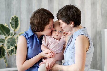 A young gay lesbian couple with a baby in their arms. Theyre posing at home. Family and adoption.
