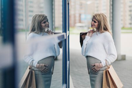 Young attractive pregnant blonde woman in a white sweater against the backdrop of the city streets. Holding a lot of shopping bags. Sunny day.の写真素材