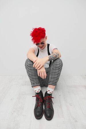 Portrait of a cool fashionable modern young girl. A short haircut with shaved temple. Dyed bright red hair. Red lipstick. Studio photo on a white background. Suspenders on checkered pants and sunglasses.の写真素材