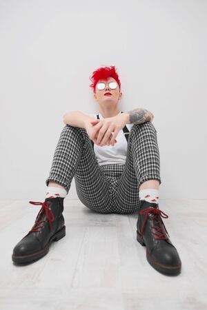 Portrait of a cool fashionable modern young girl. A short haircut with shaved temple. Dyed bright red hair. Red lipstick. Studio photo on a white background. Suspenders on checkered pants and sunglasses.の写真素材
