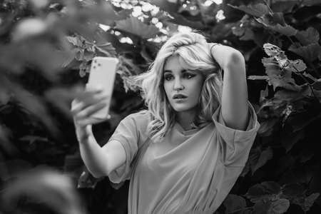 A young beautiful blonde girl is photographed on a smartphone, taking a selfie in the park against a...