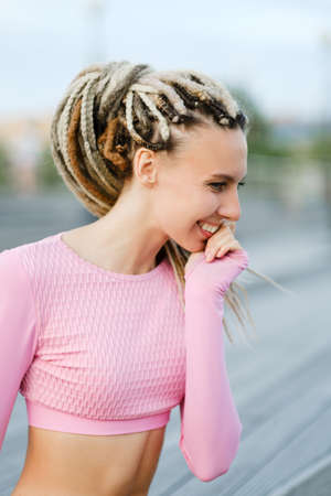 Summer urban portrait of a slender young beautiful woman with dreadlocks in a pink T-shirt and denim short shorts on a wooden staircase. The girl laugh and flirt.の写真素材